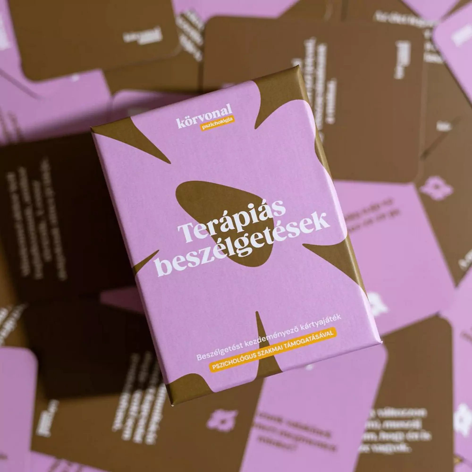 A Terápiás beszélgetések kártyajáték doboza felülnézetből, barna-rózsaszín kérdéskártyák között elhelyezve, hangulatos kompozícióban.