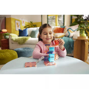 Egy mosolygó kislány játszik a MAGNA-TILES Cat Tree készlettel az asztalon. A gyerek a piros macskafigurát helyezi a színes, mágneses elemekből épített cicatorony tetejére. A háttérben otthonos nappali látható.