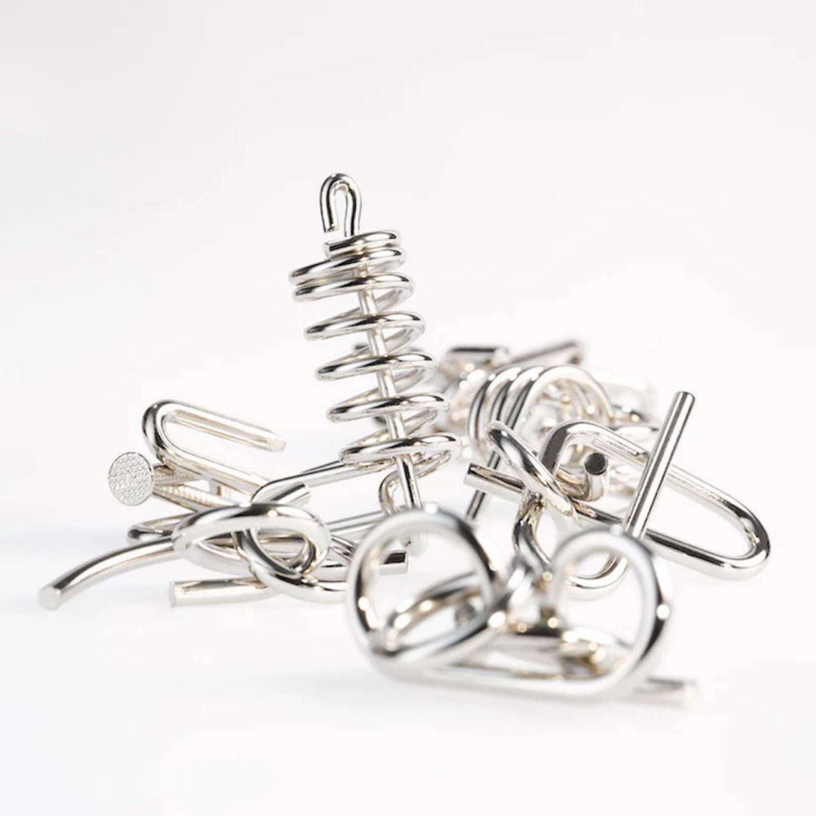 A 10 Metal Puzzle Set zöld változatának tartalma szétszedve, különböző alakú és formájú ezüstszínű fém kirakóelemek halmaza világos háttér előtt, előtérben egy spirálos forma emelkedik ki.