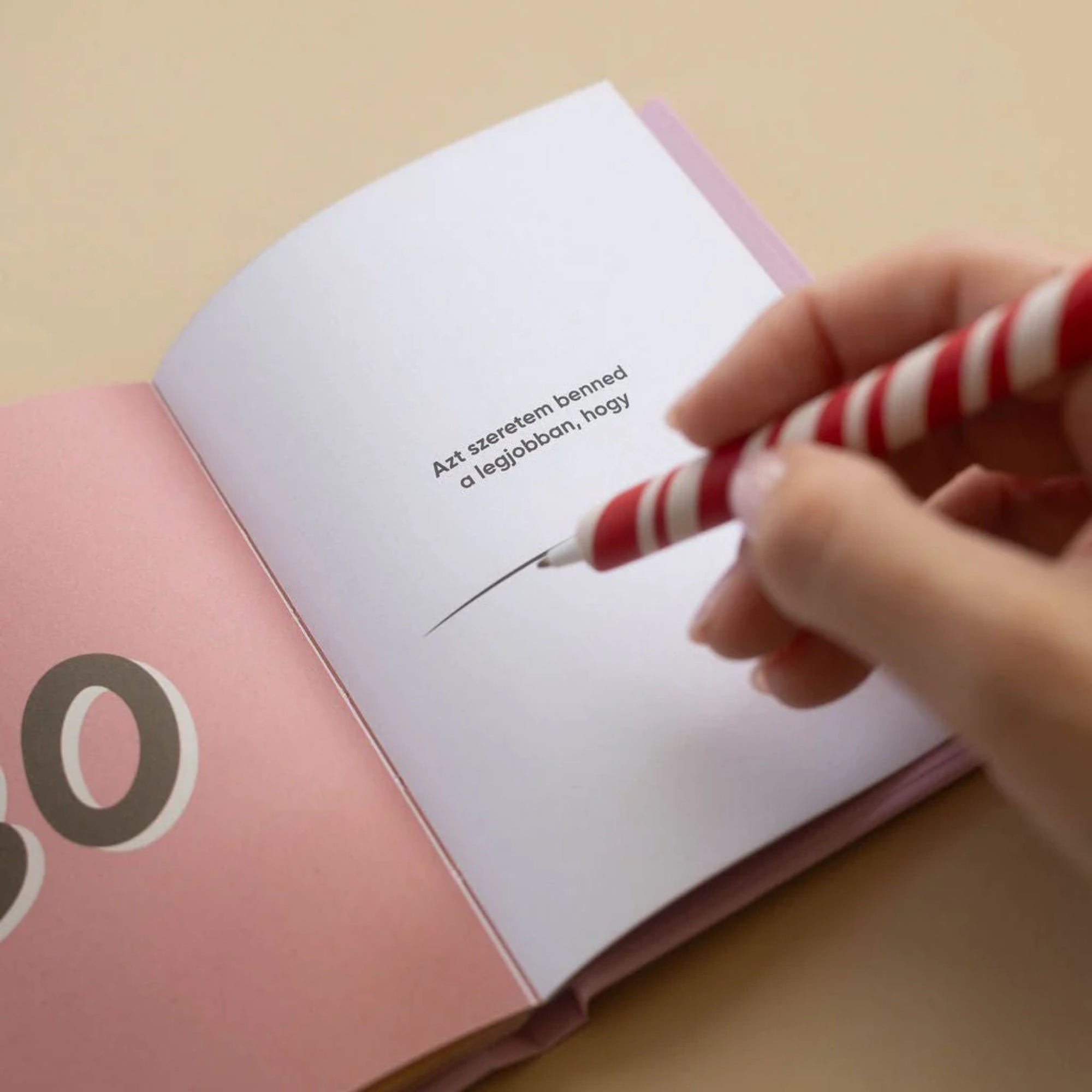 Kéz ír a „Dolgok, amiket szeretek benned, Anya” kitölthető ajándékkönyv egyik oldalára, tollal kitöltve a személyes üzenethez tartozó sort.