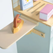 Fa vasaló egy kihúzható vasalódeszkán, mellette színes mosóporos dobozok és kiegészítők, pasztell árnyalatú mosoda felületen.