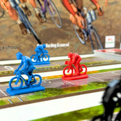 Flamme Rouge társasjáték piros és kék színű kerékpáros figurái a versenypályán, részletesen kidolgozott biciklis alakokkal és nyomtatott útszakasz elemekkel, játék közbeni helyzet bemutatása.