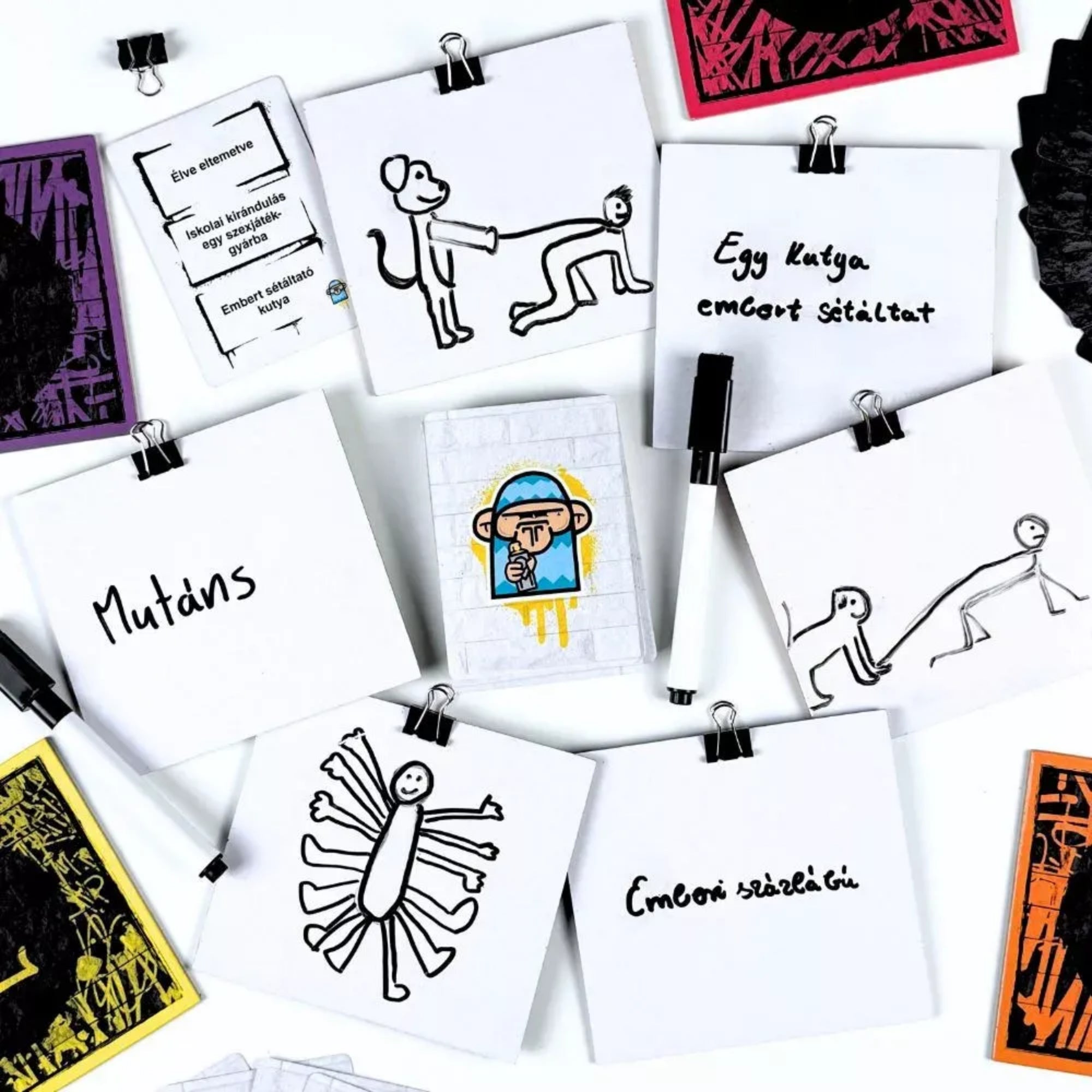 G.E.C. – Skicc játék közben: rajzlapokra készült egyszerű, humoros ábrák és feliratok, középen a feladatkártyával, körülötte filcek és csipeszek.