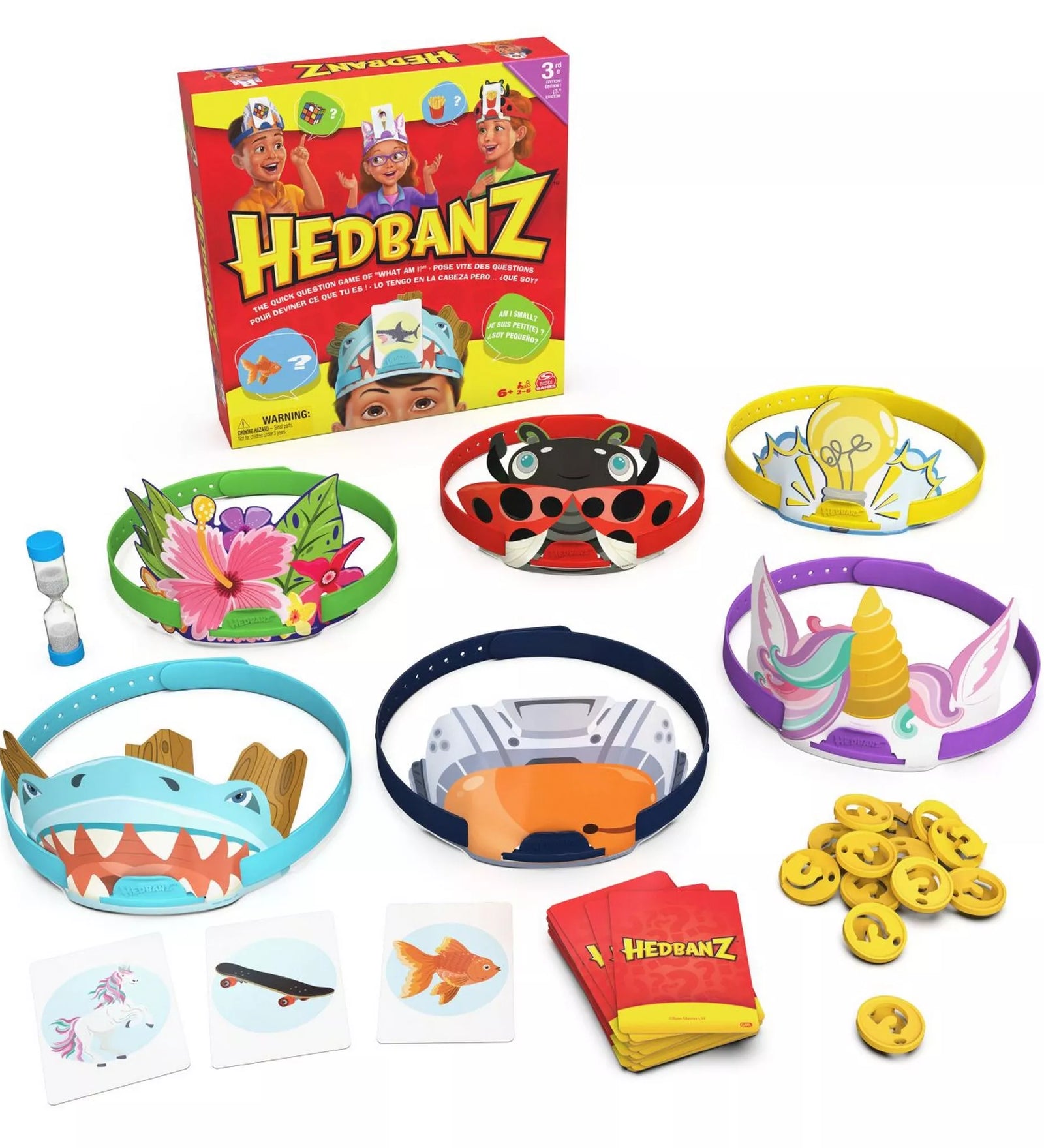 A Hedbanz társasjáték tartozékai: hat különböző fejpánt (cápa, unikornis, katica, villanykörte, virág, űrhajós), kártyák, homokóra és sárga jelzőzsetonok, a háttérben a játéktartó dobozzal.