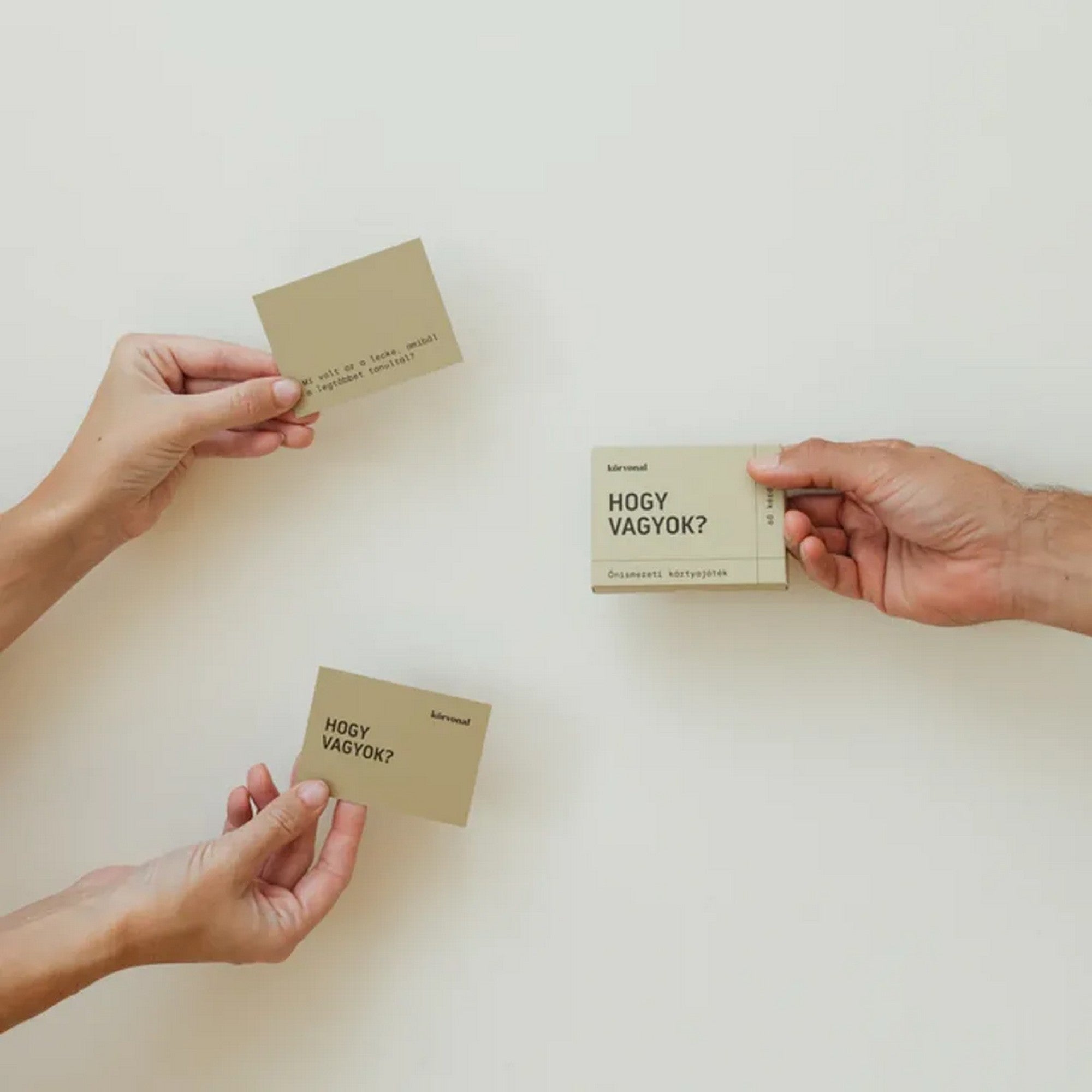 Három kéz tartja a Körvonal – Hogy vagyok? önismereti kártyajáték lapjait és a dobozt, világos háttér előtt.