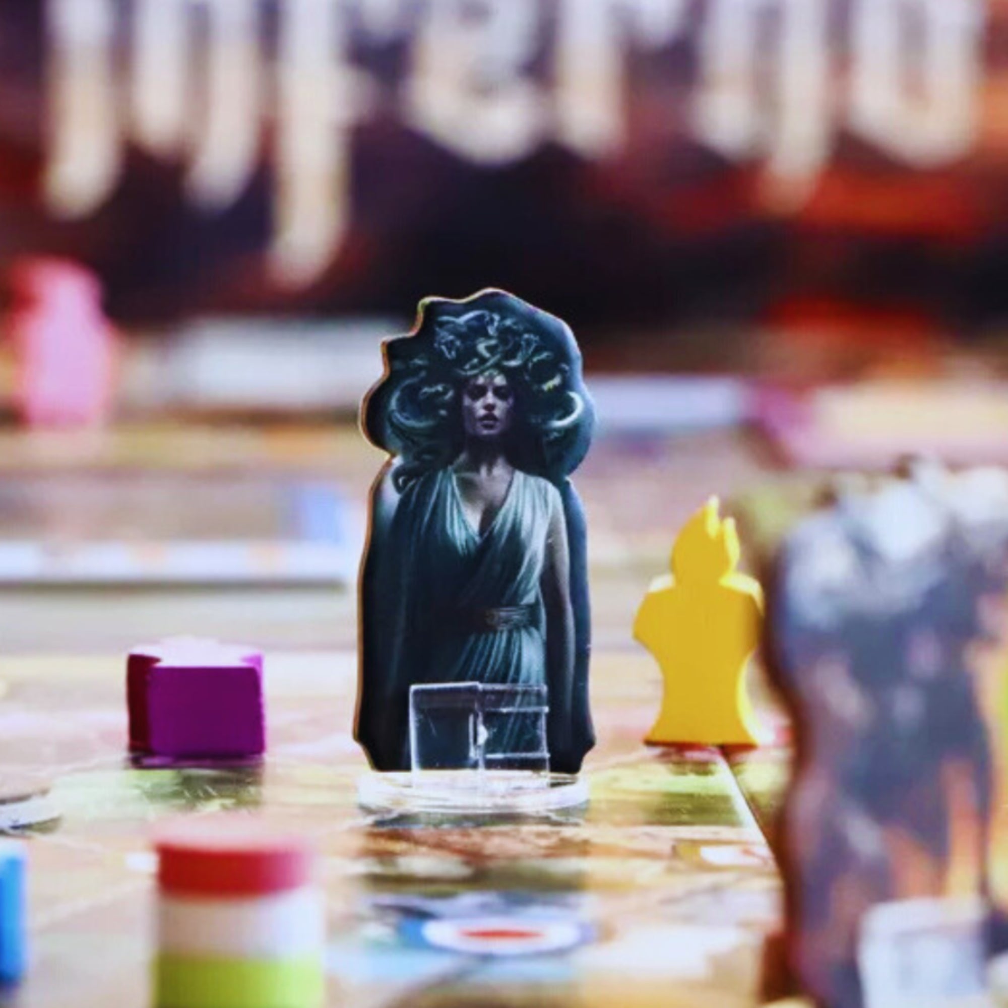 A Inferno társasjáték egyik figurája, egy Medúzát ábrázoló álló báb, színes játékelemek és kártyák között, részletes és hangulatos asztali jelenetben.