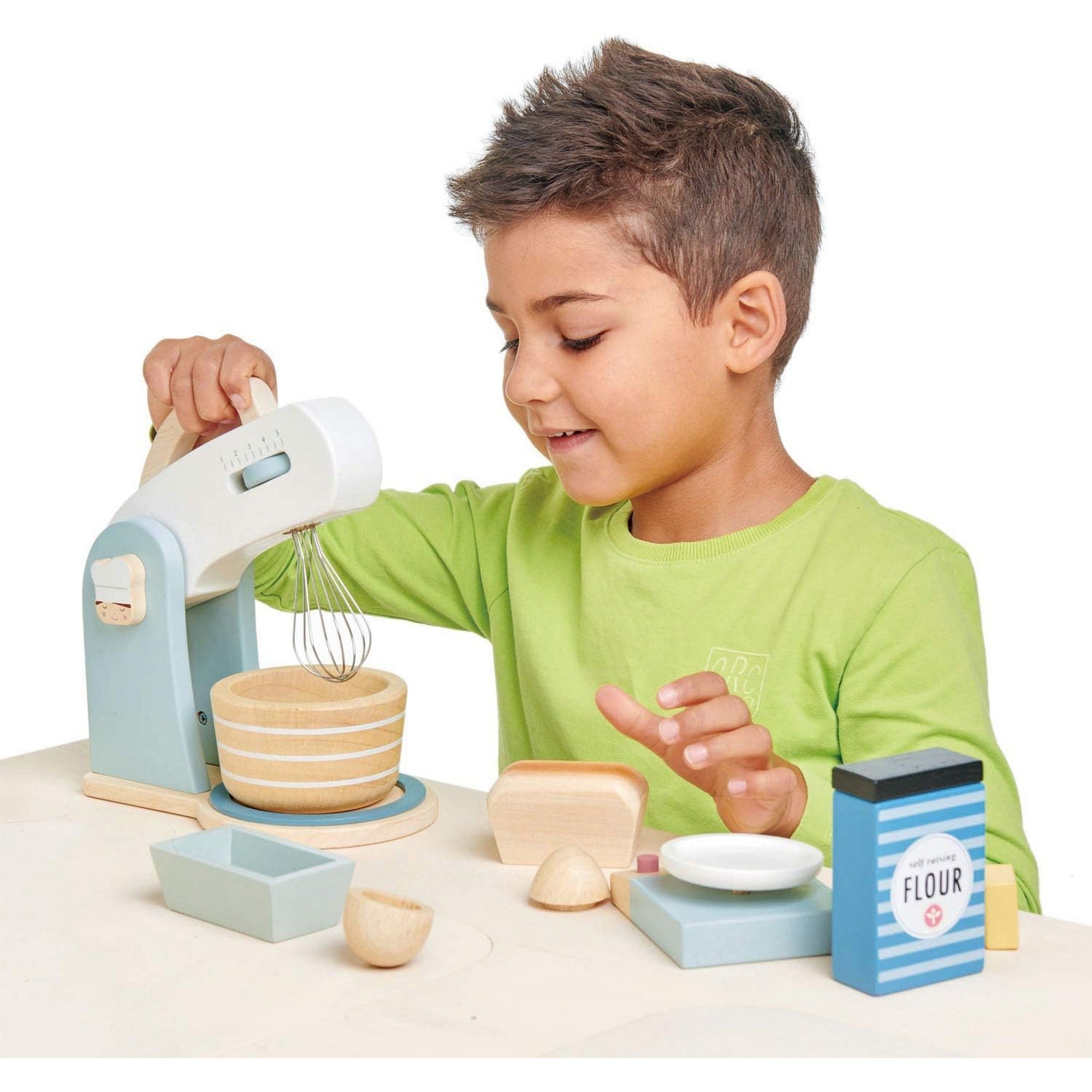 Harmadik kép: Egy kisfiú játszik a Tender Leaf Toys Mini Chef fa sütőkészlettel, a habverőt forgatja, mellette a játék alapanyagok sorakoznak.