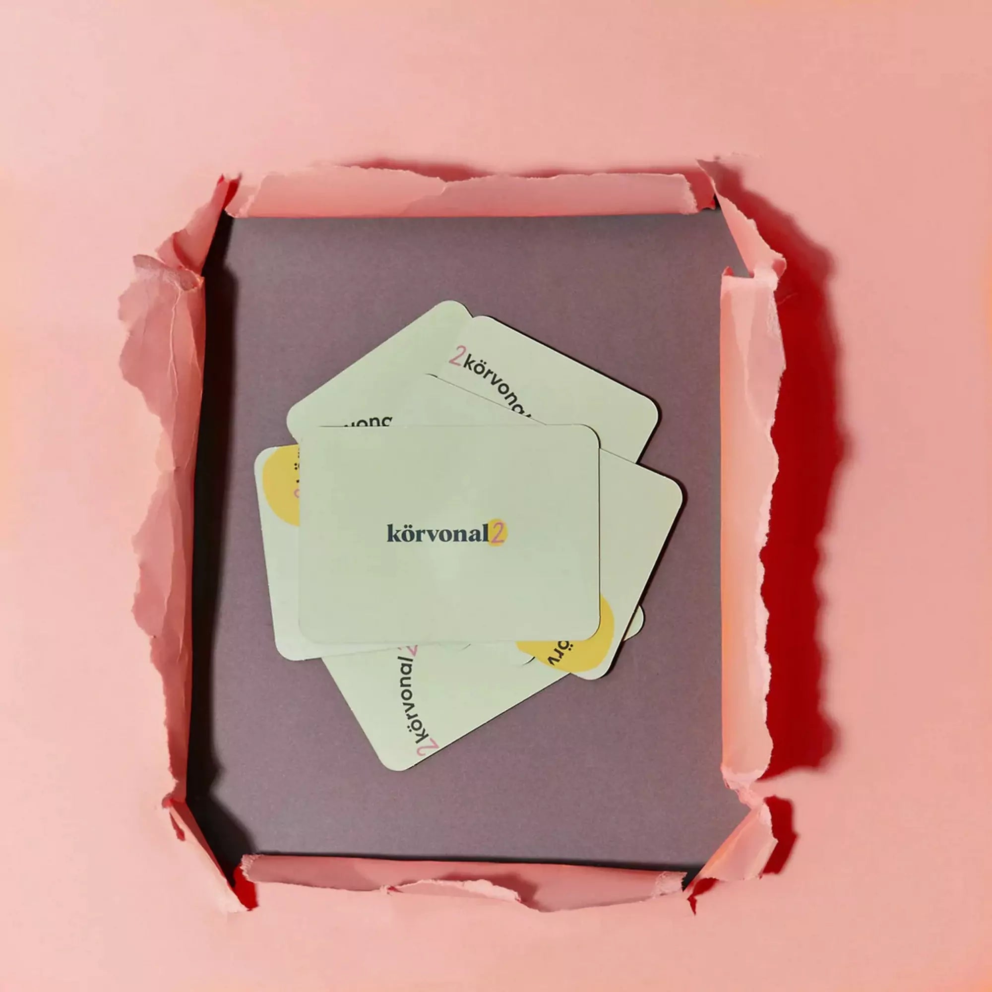 Kicsomagolt Körvonal 2 kártyajáték pasztellrózsaszín háttérrel, néhány arccal lefelé fordított kártyával, középen a logóval ellátott lappal.