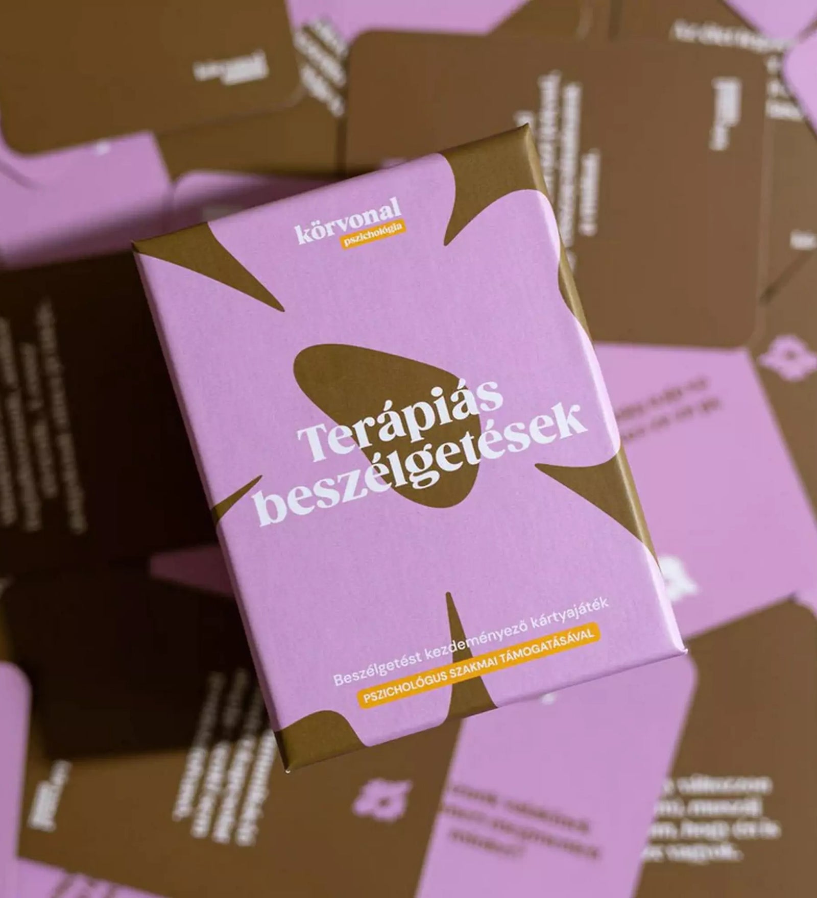 A Terápiás beszélgetések kártyajáték doboza felülnézetből, barna-rózsaszín kérdéskártyák között elhelyezve, hangulatos kompozícióban.