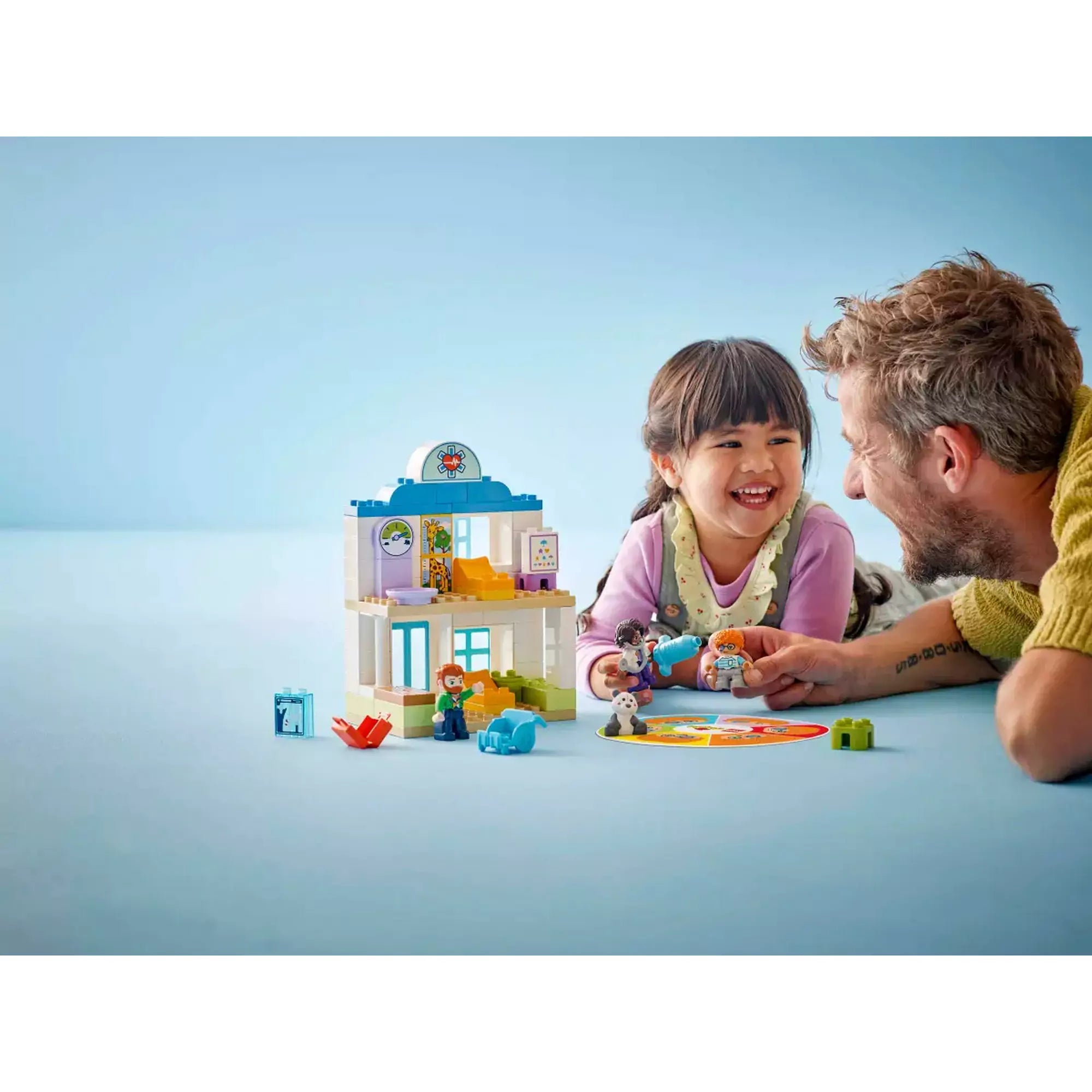 Egy apuka és kislánya közösen játszanak a LEGO DUPLO Első látogatás az orvosnál (10449) készlettel, amely barátságos, orvosi jelenetet ábrázol.