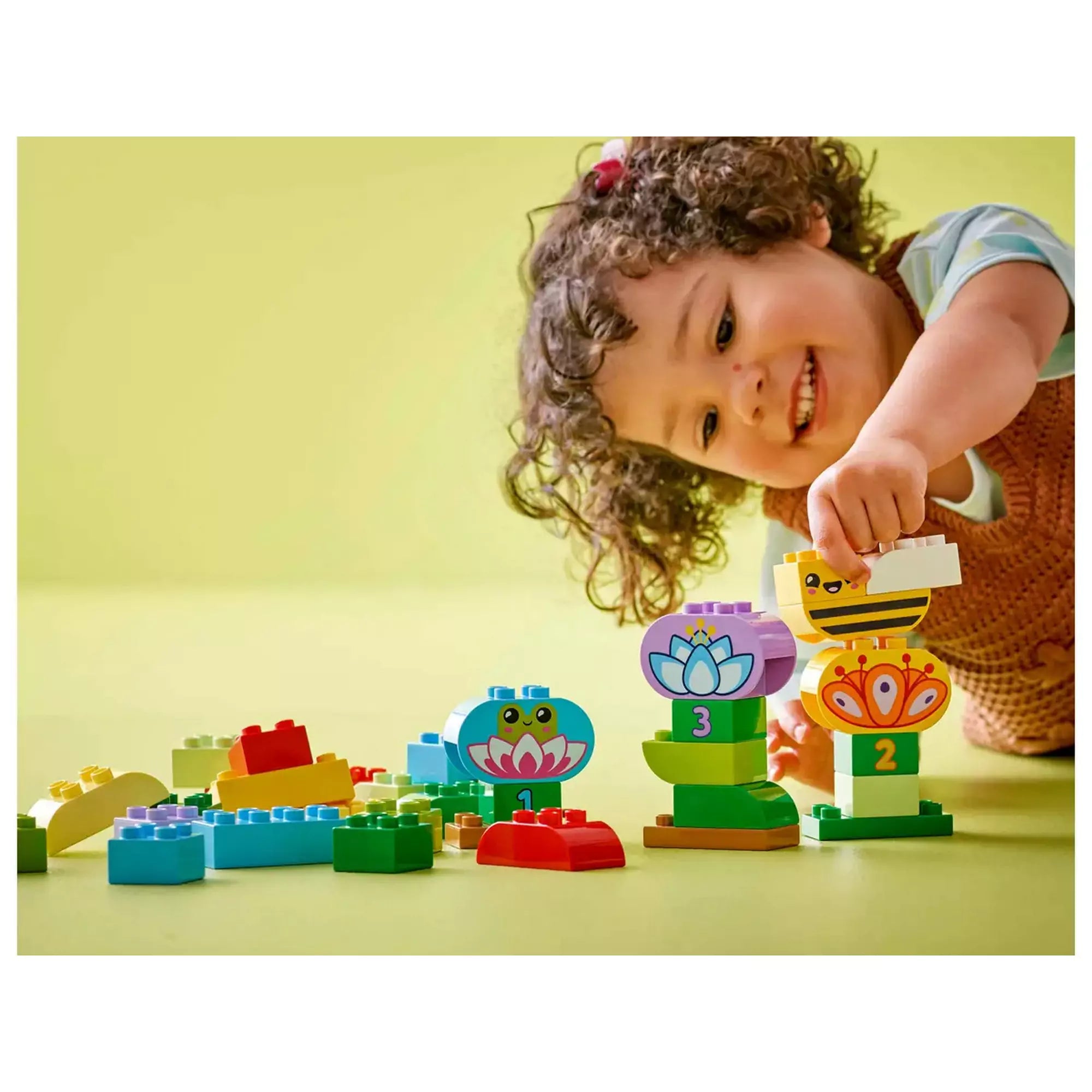 Egy mosolygó kisgyermek játszik a LEGO DUPLO Kreatív kert és virágok (10444) színes elemeivel, amelyek között méhecske, béka és pillangó is megtalálható.

