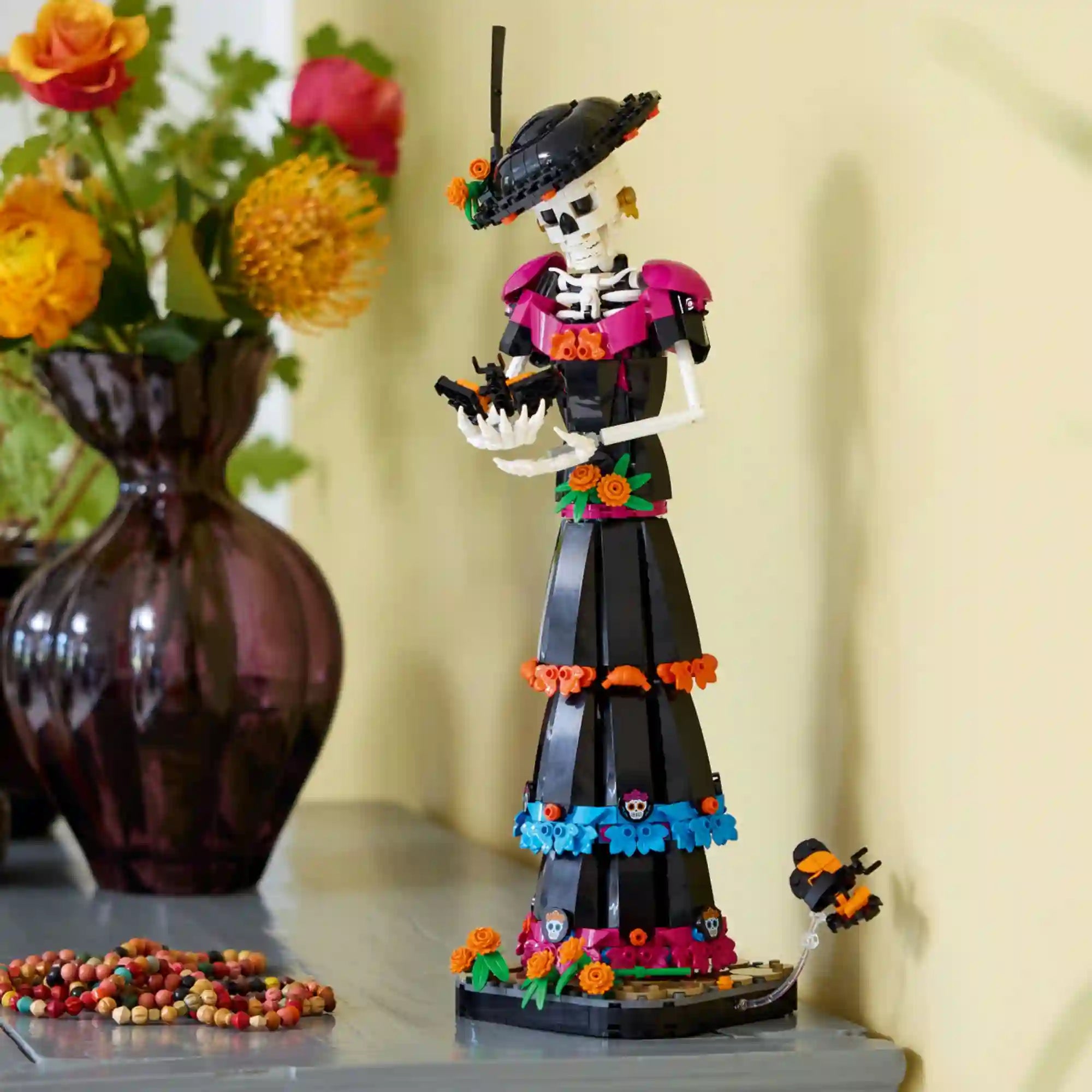 A LEGO Ideas La Catrina 21372 megépített La Catrina figurája dekorációként egy polcon, virágokkal és méhecskével.