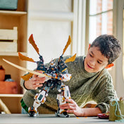 Fiú játszik a LEGO NINJAGO Cole titánsárkány-robotja (71821) összeépített modellel egy beltéri asztalon, jól láthatóak az arany pengék és a mozgatható végtagok.