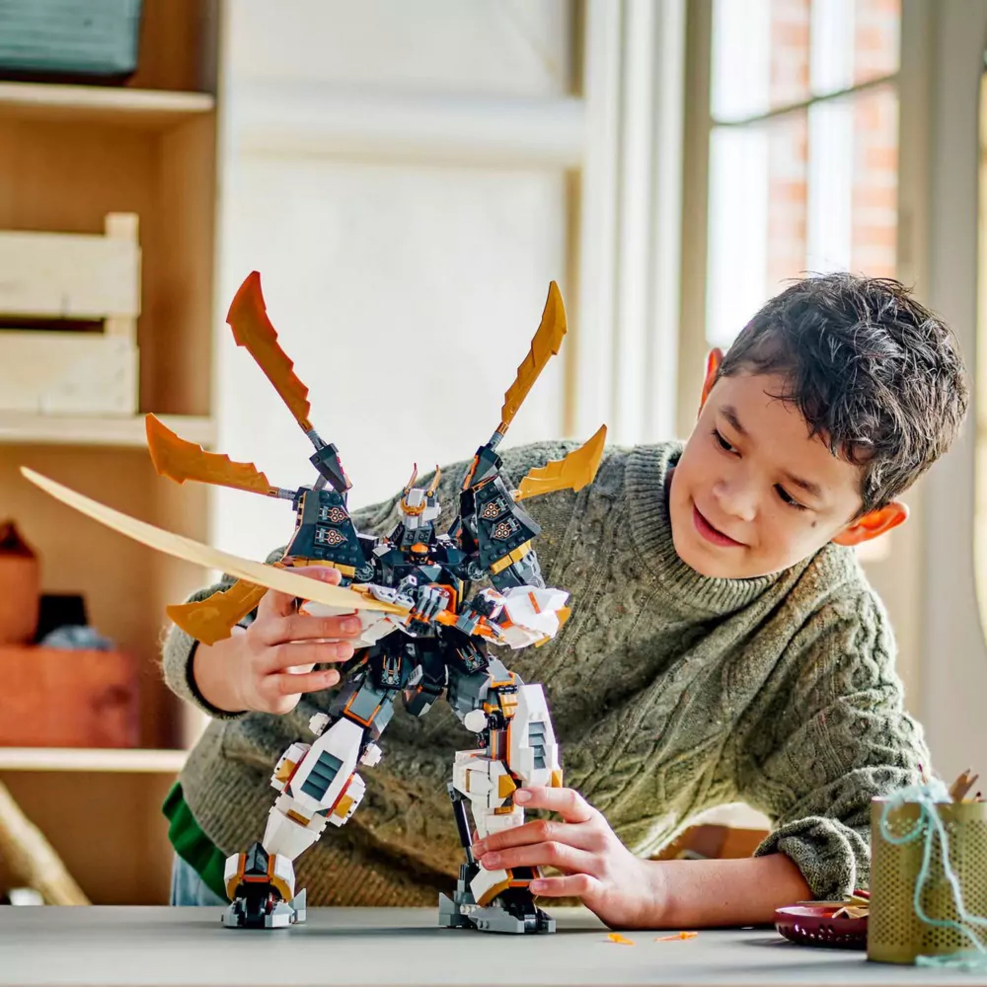 Fiú játszik a LEGO NINJAGO Cole titánsárkány-robotja (71821) összeépített modellel egy beltéri asztalon, jól láthatóak az arany pengék és a mozgatható végtagok.