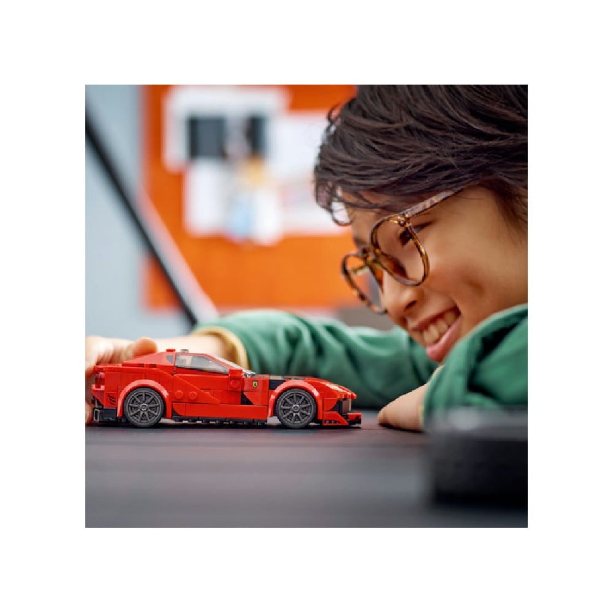 Egy gyerek játszik a LEGO Ferrari 812 Competizione modellel, mosolyogva az asztalon tologatja az autót, háttérben homályos szobaelemek.