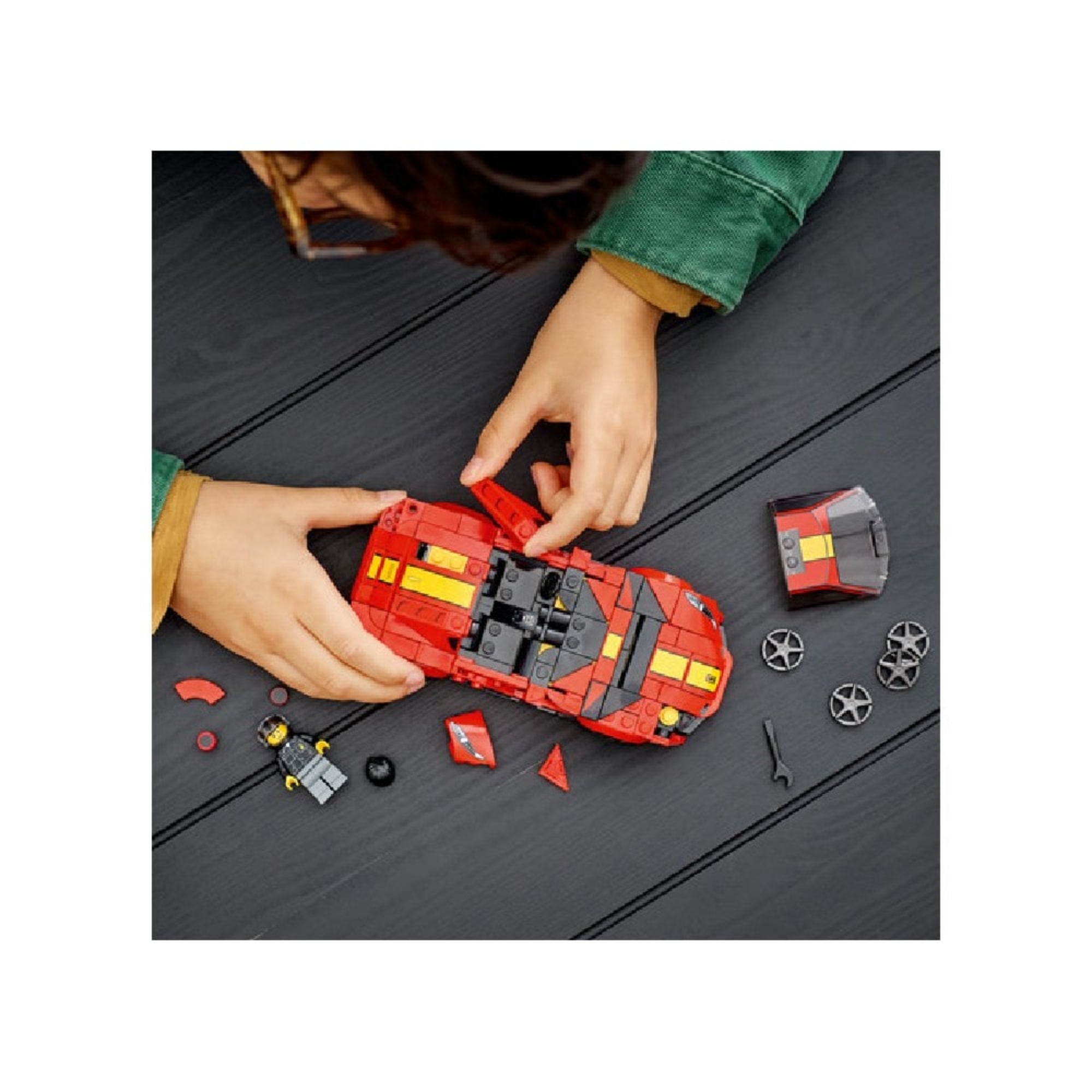 Felülnézeti kép egy gyermek kezéről, aki éppen a LEGO Ferrari 812 Competizione-t építi, körülötte kockák, kiegészítők és minifigura.
