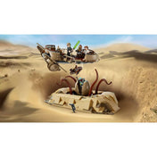 Akciódús illusztráció a LEGO Star Wars sivatagi jelenetéről. A sarlacc üregéből előtörő csápok épp elkapni próbálják az egyik zuhanó minifigurát, miközben a sikló lebegve halad el fölötte. A háttérben a homokdűnék végtelenje és a kék égbolt látható.