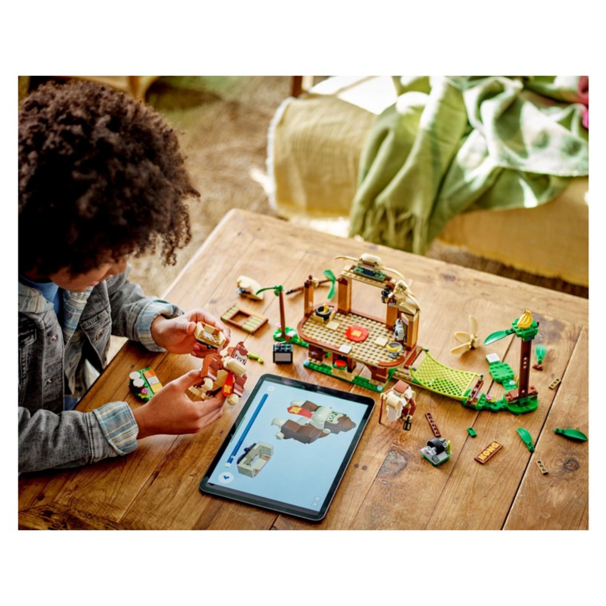 Egy gyermek LEGO Super Mario Donkey Kong lombháza kiegészítő szettjét (71424) rakja össze egy asztalon, miközben egy tablet digitális útmutatót jelenít meg a Donkey Kong figurához.