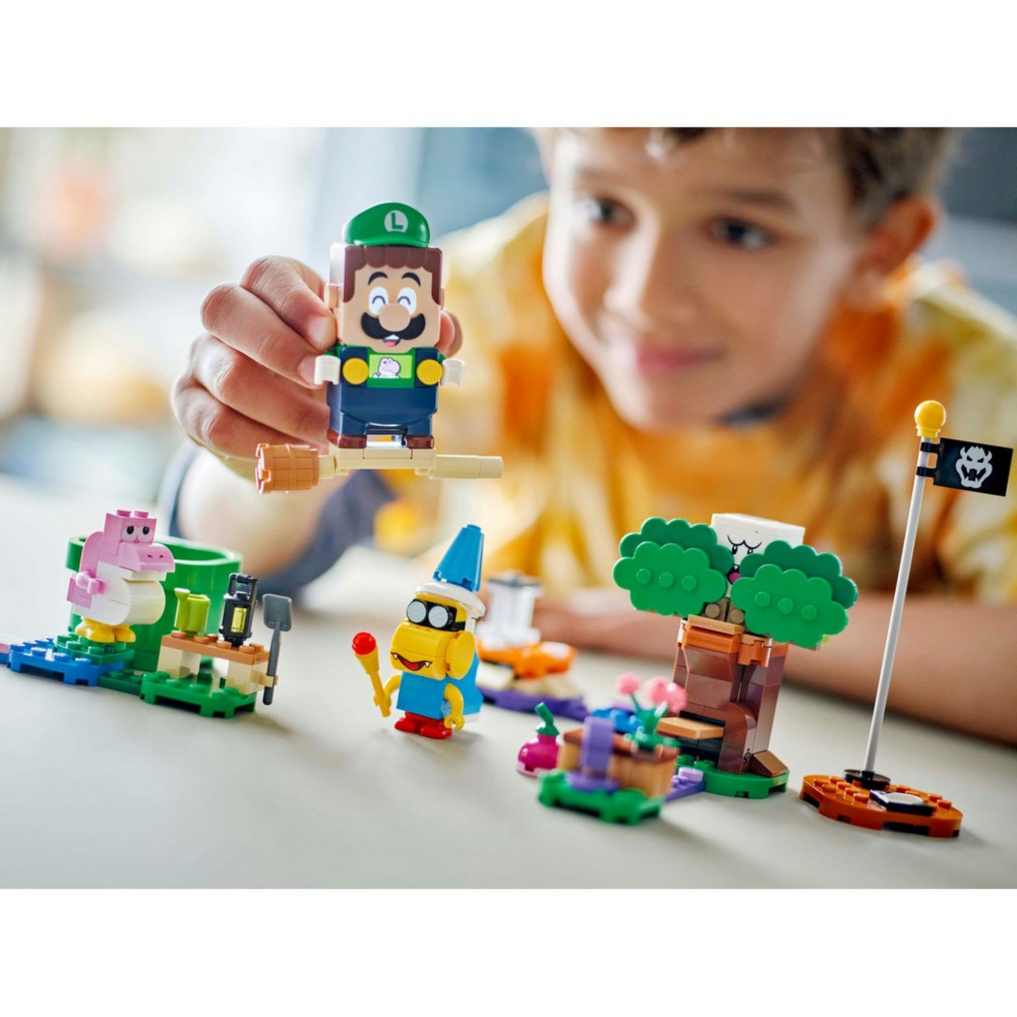 Egy kisfiú izgatottan emeli fel a LEGO Luigi figurát, miközben játszik a LEGO Super Mario 71440 készlet színes karaktereivel és építményeivel, köztük Baby Yoshival és Kamekkel.