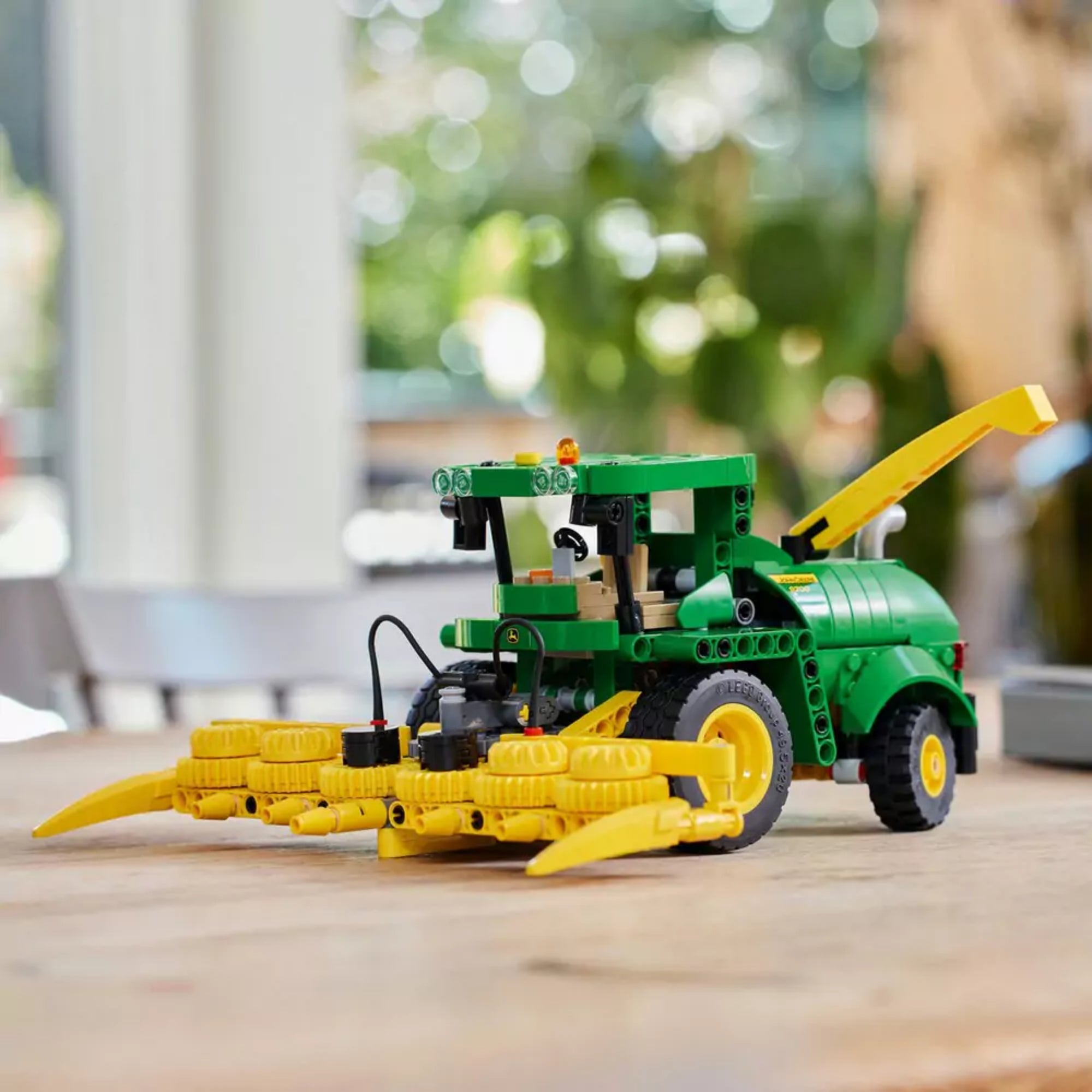 Összeépített John Deere 9700 LEGO Technic modell természetes asztali környezetben, háttérben elmosódott beltéri növényzettel és fénypontokkal.