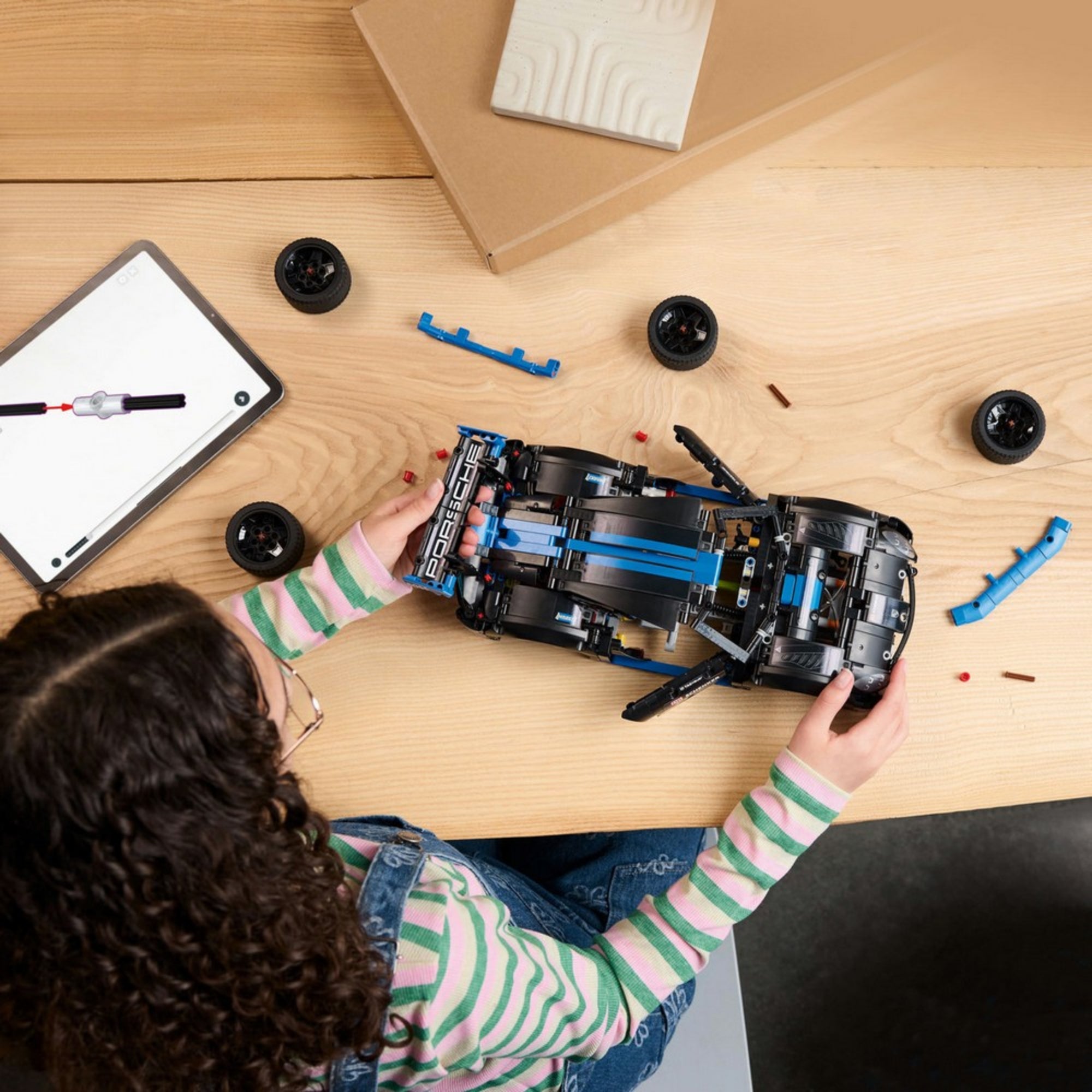 Egy fiatal lány építi a LEGO Technic Porsche GT4 e-Performance versenyautót, az asztalon szétszedett kerekek és alkatrészek, mellette egy tablet digitális útmutatóval.