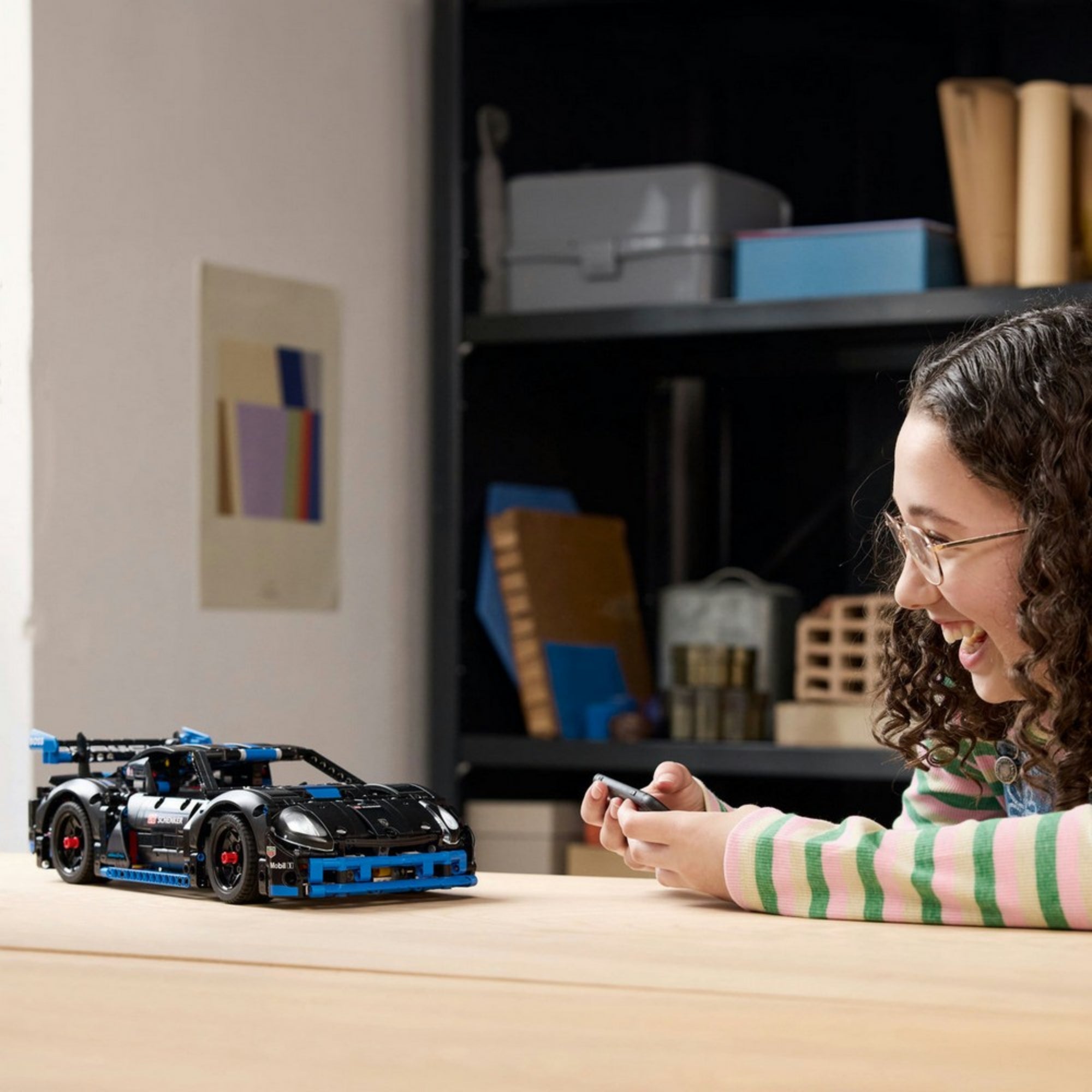 Egy lány mosolyogva irányítja a LEGO Technic Porsche GT4 e-Performance versenyautót távirányítóval, az autó lendületesen halad az asztalon.