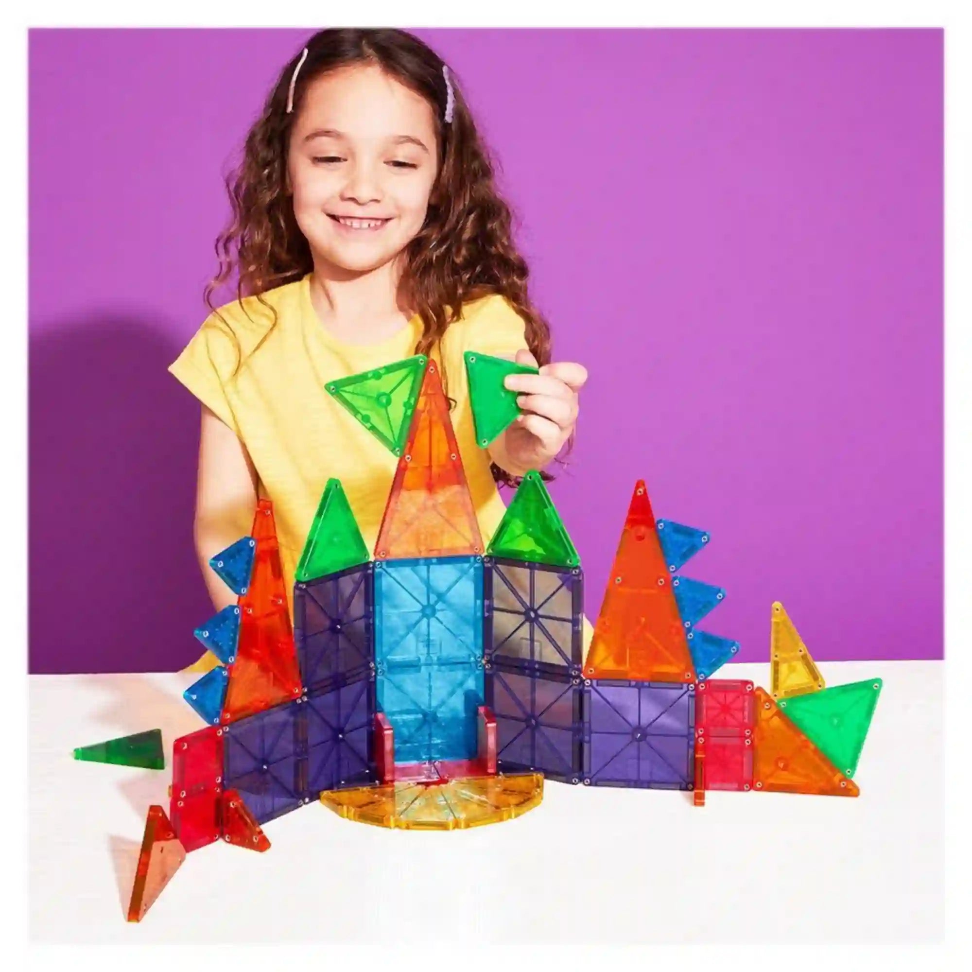 Mosolygó kislány sárga pólóban, amint a MAGNA-TILES Combo microMAGS 46 darabos építőkészlet színes mágneses elemeiből készült kastélyhoz zöld háromszöget illeszt.