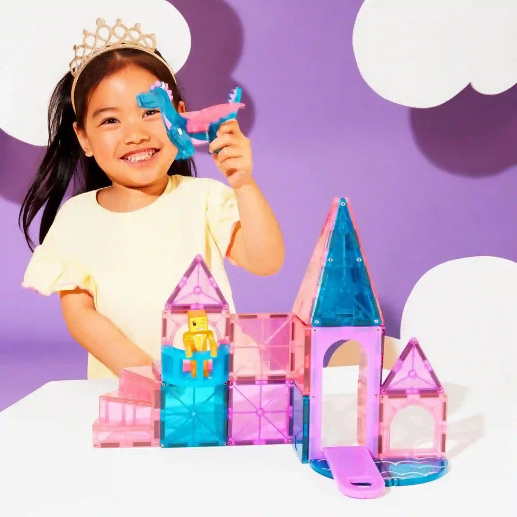 Mosolygó, koronát viselő kislány játszik a MAGNA-TILES Castle 25 darabos építőkészlettel, kezében kék-rózsaszín sárkányfigurát tart a megépített kastély fölött.