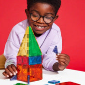 Mosolygó, szemüveges kisfiú épít a MAGNA-TILES Combo + microMAGS 20 darabos mágneses építőkészlettel, kezében lila háromszög alakú mágneses lapot tart.