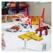 MAGNA-TILES Farm Animals mágneses építőkészlet farmjelenete, amelyben egy gyermek fehér tyúkfigurát tart a foltos tehén fölé, a barna ló és szénabálák mellett.