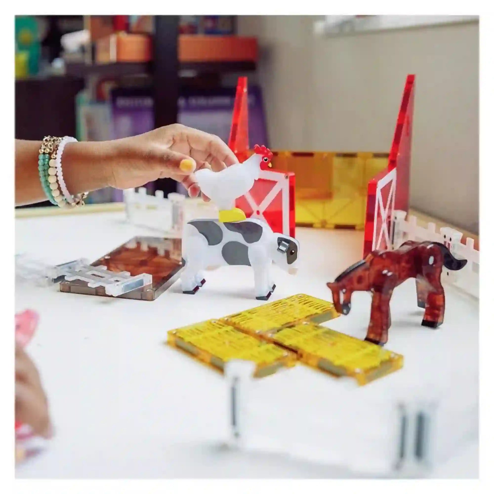 MAGNA-TILES Farm Animals mágneses építőkészlet farmjelenete, amelyben egy gyermek fehér tyúkfigurát tart a foltos tehén fölé, a barna ló és szénabálák mellett.