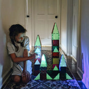 Egy kislány sötét folyosón játszik a Magna-Tiles Glow készlettel, egy világító kastélyt építve. Az elemek foszforeszkáló hatása lenyűgöző látványt nyújt.