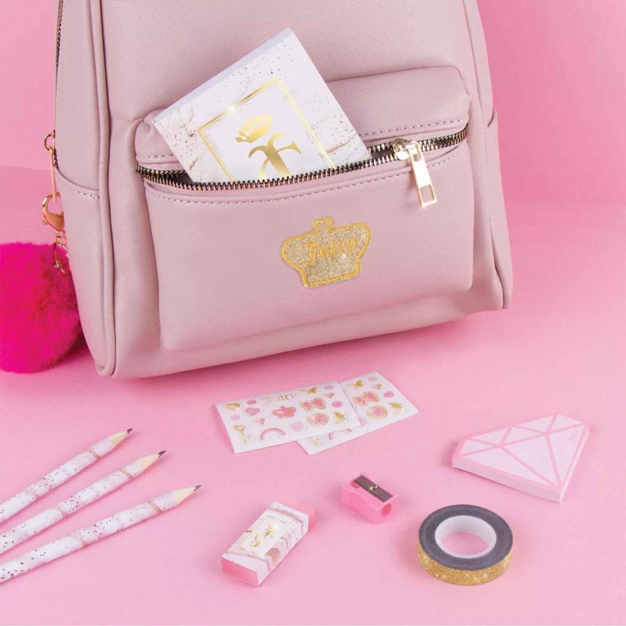 Juicy Couture Deluxe Stationery Set elemei rózsaszín háttéren, füzet a táskában, ceruzák, radír, hegyező, matricalap és arany dekortapasz elrendezve.