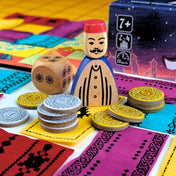 Marrakech társasjáték részletfotó a fa piacktulajdonos figuráról, dobókockáról és arany-ezüst pénzérmékről a szőnyeglapkák között.