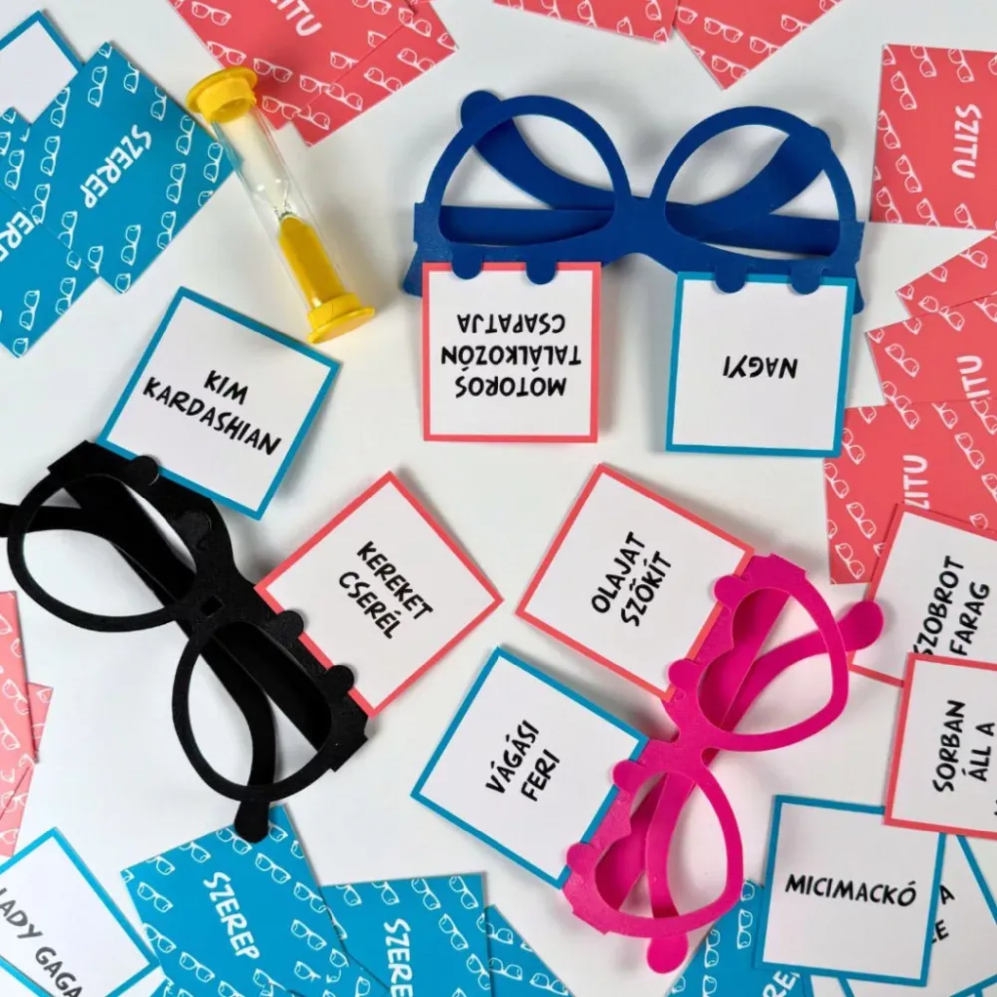 Nagyi bugyiban társasjáték játékelemei közelről: színes műanyag szemüvegekbe helyezett szerep- és szitukártyák, feliratos kártyák és homokóra játék közbeni elrendezésben.