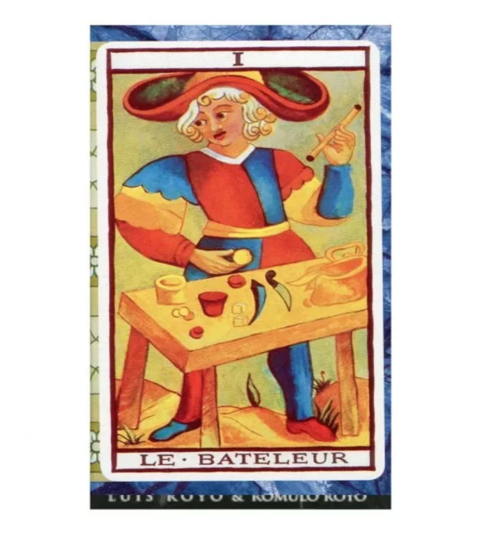 A képen egy színes, klasszikus stílusban illusztrált tarot kártyalap látható, amely „Le Bateleur” (A Mágus) nevet viseli. A figura vörös-kék-sárga ruhát visel, fején széles karimájú kalappal, és egy pálcát tart a kezében. Előtte egy asztal látható különféle tárgyakkal – serleg, pénzérmék, kard –, amelyek a tarot négy szimbólumát jelképezik. A háttér élénk színei és a rajz stílusa a Marseille-i tarot hagyományait idézi.