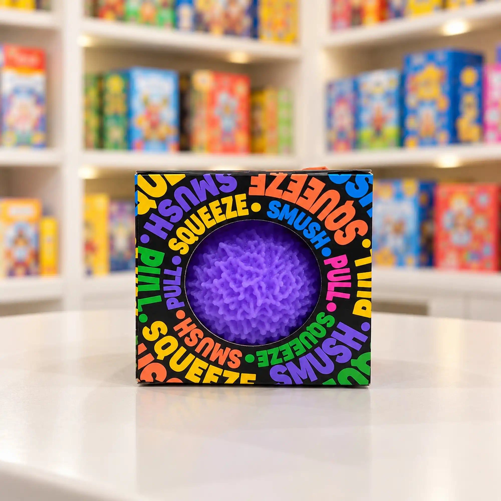 Cool Mix Squishy bolyhos tapintású labda 6 cm stresszlevezető játék (lila), fekete, színes feliratokkal díszített dobozban, kör alakú ablakon keresztül látható.