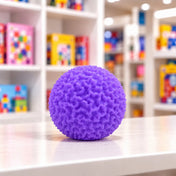 Cool Mix Squishy: Bolyhos tapintású labda 6 cm stresszlevezető játék (lila), rücskös felületű lila labda játékbolt pultján, elmosódott polcok előtt.