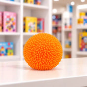 Cool Mix bolyhos tapintású squishy stresszlabda (narancssárga) – élénk narancssárga, puha tüskékkel borított, gömb alakú stresszlabda fehér asztalon, elmosódott játékbolti háttérrel.