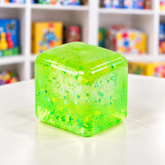 Cool Mix Squishy: Super Gel Cube stresszlevezető játék (zöld) – Átlátszó, élénkzöld, csillámos gélkocka lekerekített sarkokkal, fehér asztalon, elmosódott játékbolti háttér előtt.