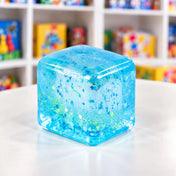 Cool Mix Squishy: Super Gel Cube stresszlevezető játék (kék) – Átlátszó, kék, csillámos gélkocka lekerekített sarkokkal, fehér asztalon, elmosódott játékbolti háttér előtt.