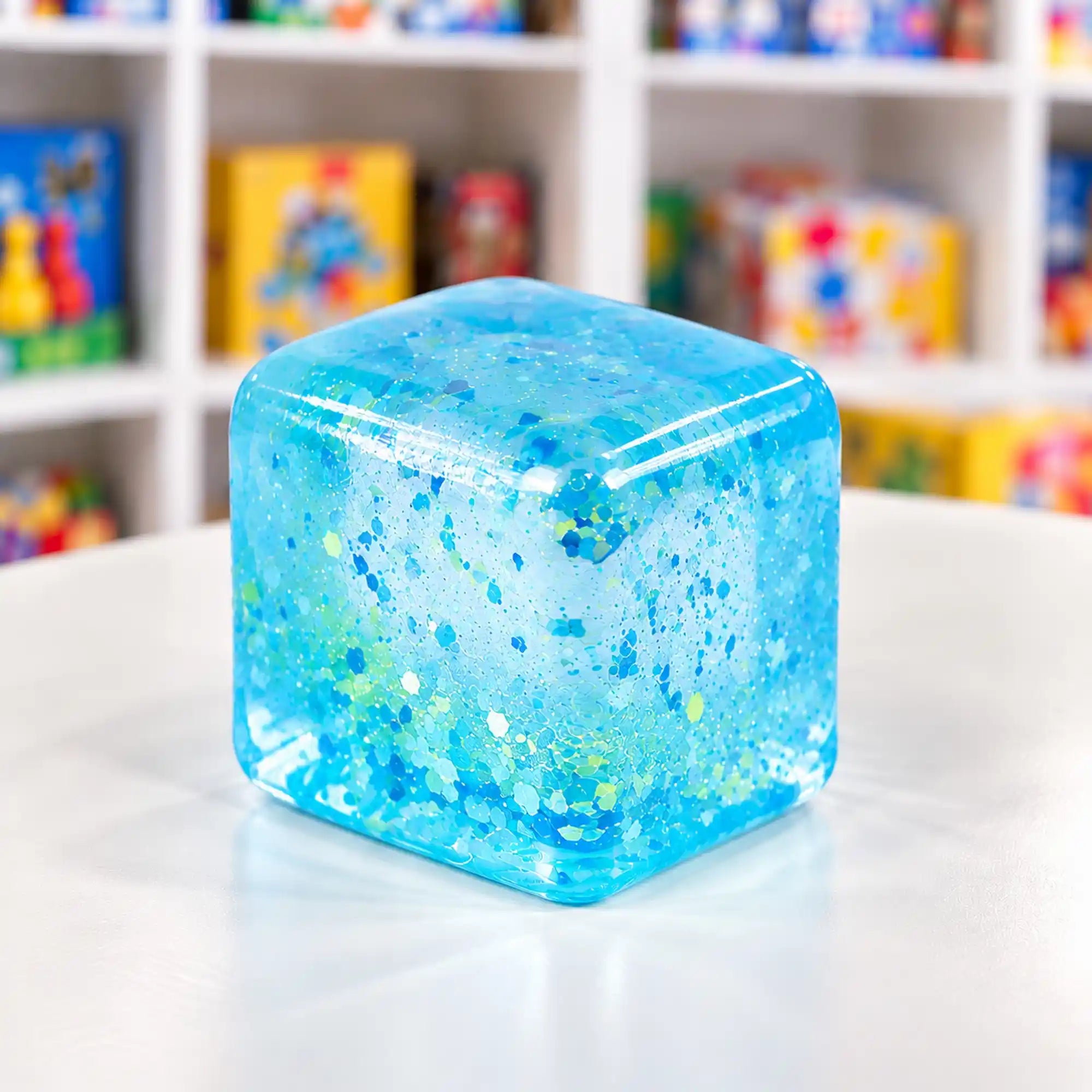 Cool Mix Squishy: Super Gel Cube stresszlevezető játék (kék) – Átlátszó, kék, csillámos gélkocka lekerekített sarkokkal, fehér asztalon, elmosódott játékbolti háttér előtt.