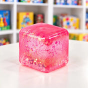 Cool Mix Squishy: Super Gel Cube stresszlevezető játék (rózsaszín), áttetsző, csillámos, lekerekített sarkú zselékocka fehér asztalon.