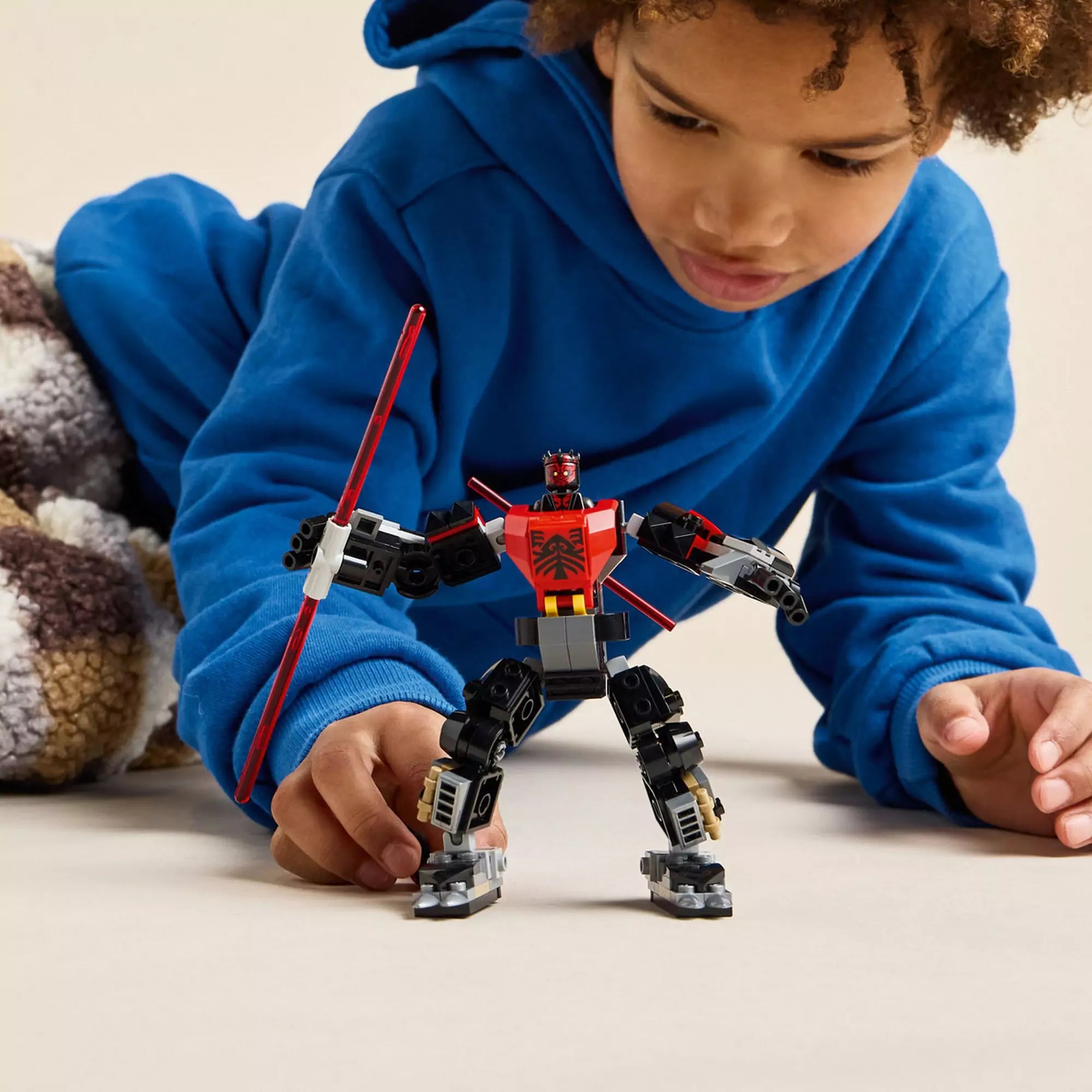 Egy kisfiú figyelmesen játszik a teljesen megépített Darth Maul mecha figurával, amely vörös fénykardot tart a kezében.