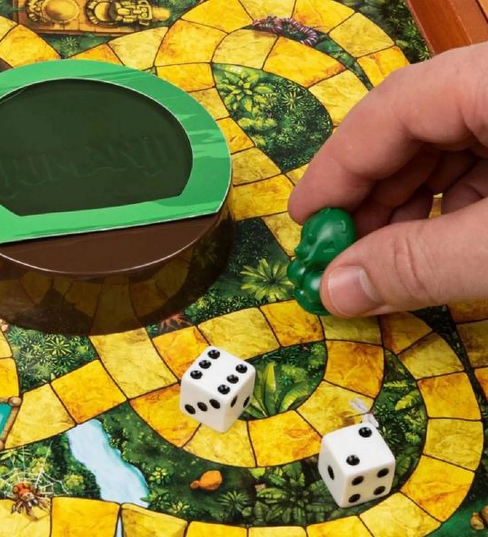 Közelkép a Jumanji társasjáték játéktáblájáról, amely dzsungel mintázatú, sárga ösvényekkel. Egy játékos keze zöld bábut helyez a pályára a dobókockák mellé, a középen található zöld Jumanji kupola mellett.