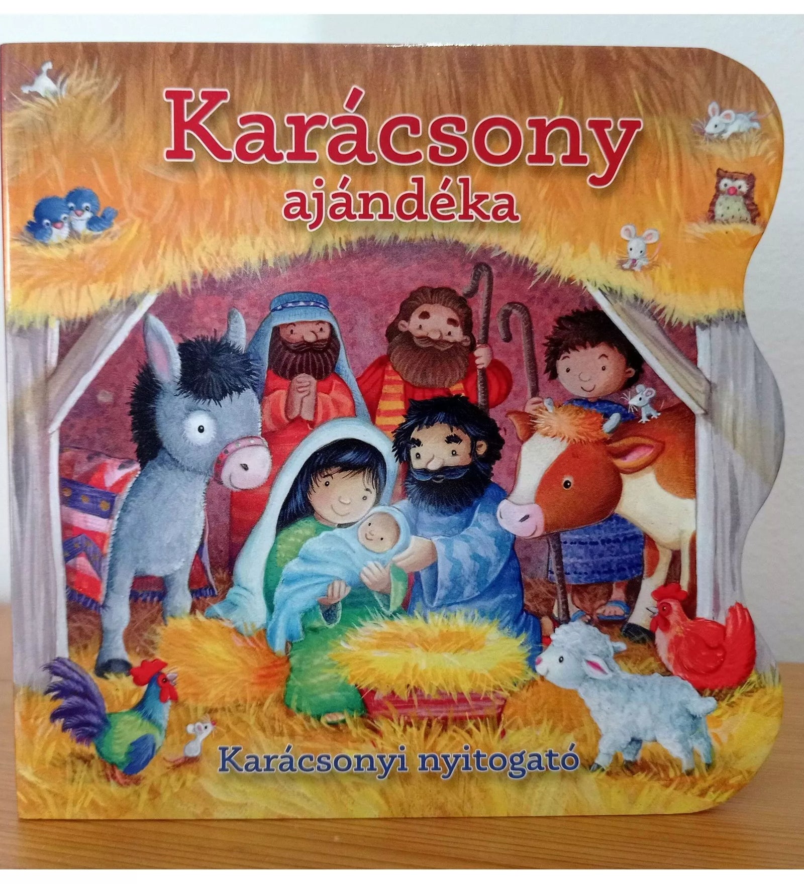 A Karácsony ajándéka – Karácsonyi nyitogató keménylapos könyv borítója, amelyen a betlehemi jelenet látható: Mária és József a kis Jézussal, körülöttük pásztorok és állatok vidám, színes illusztrációval.