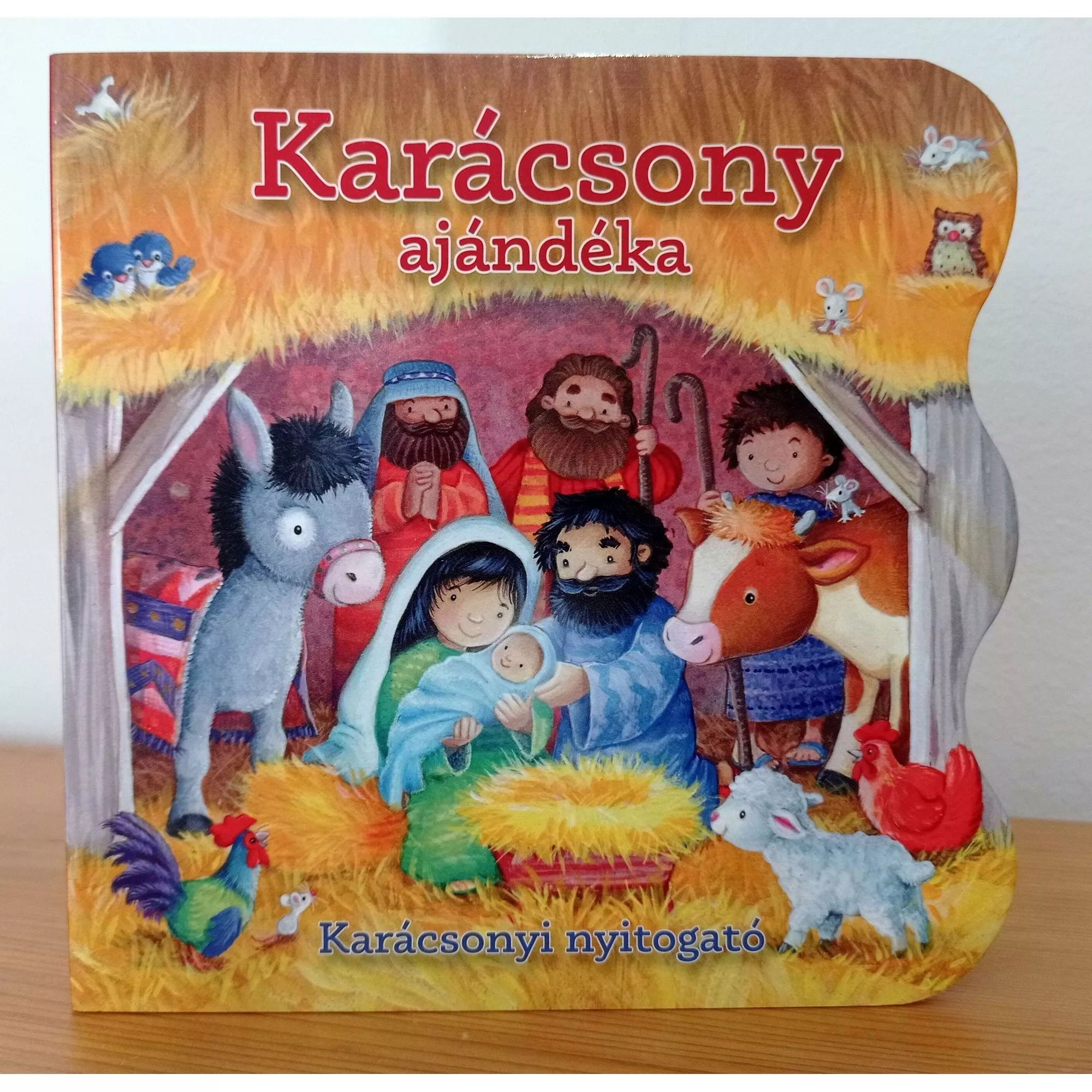 A Karácsony ajándéka – Karácsonyi nyitogató keménylapos könyv borítója, amelyen a betlehemi jelenet látható: Mária és József a kis Jézussal, körülöttük pásztorok és állatok vidám, színes illusztrációval.