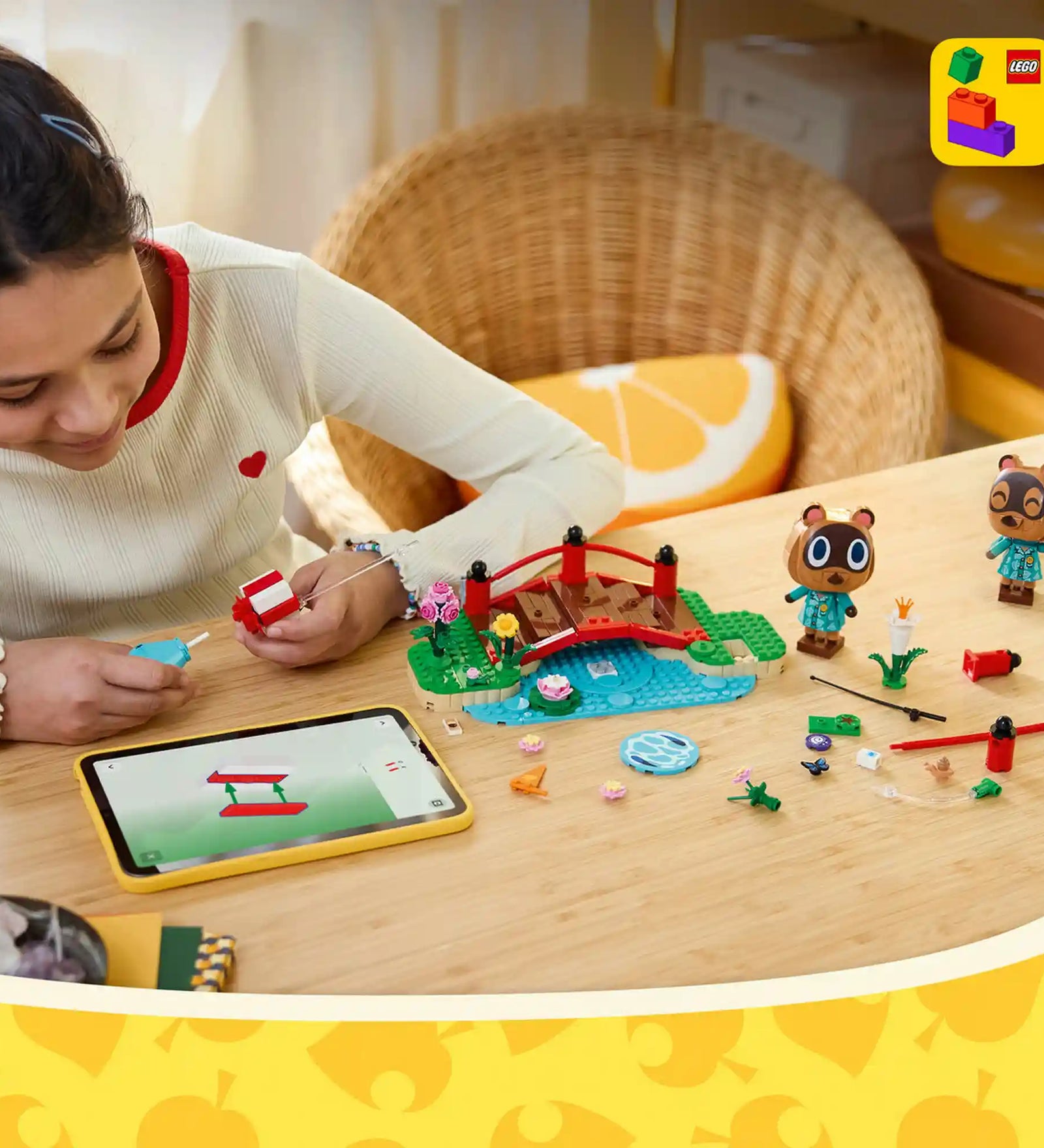 A LEGO Animal Crossing: Timmy és Tommy vidám kirándulása 77059 készlettel egy gyermek építi a piros hidat és a tóparti jelenetet a LEGO Builder alkalmazás útmutatóját követve, miközben Timmy és Tommy figurái, valamint a kiegészítők az asztalon sorakoznak.