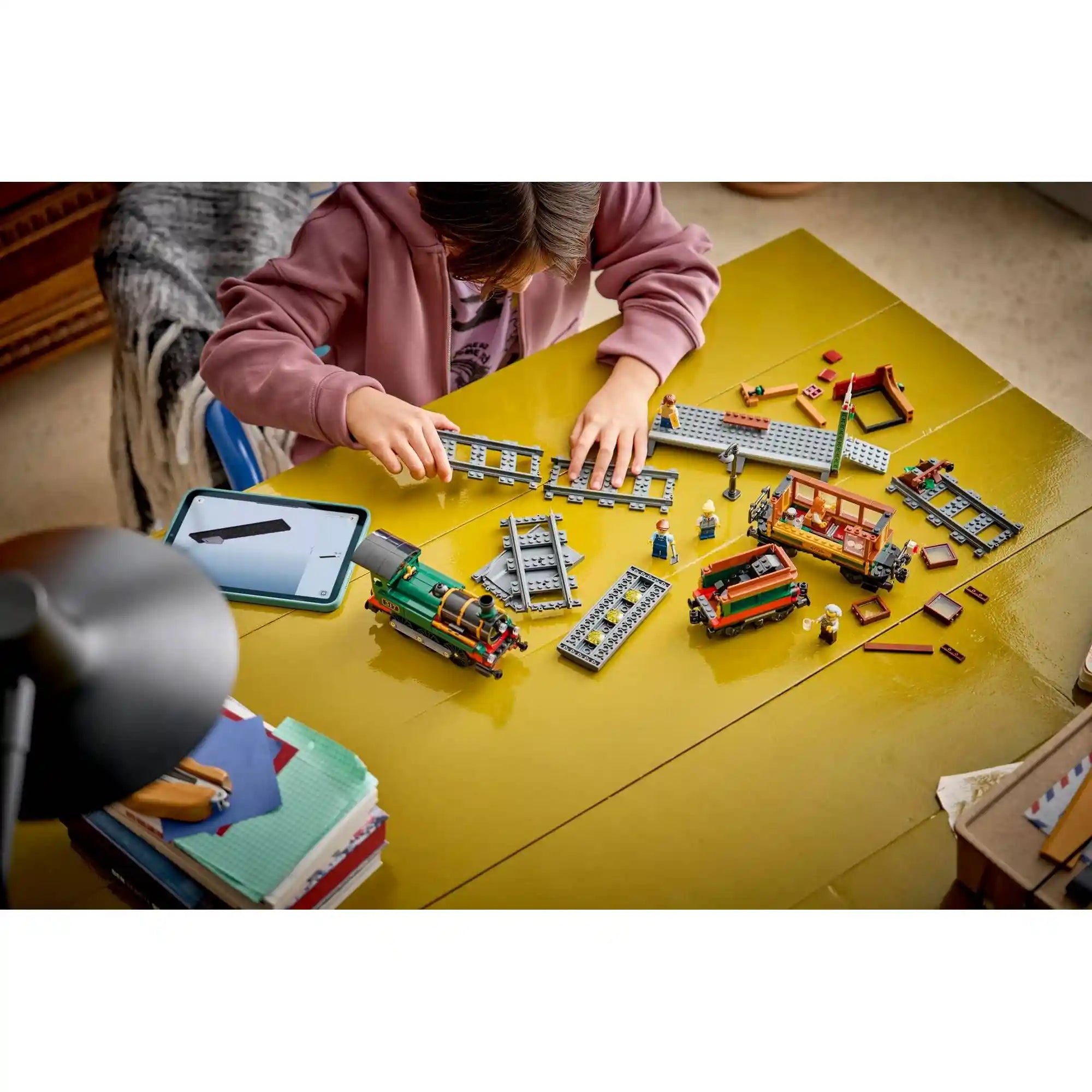 Gyermek építi a LEGO City Veterán gőzvonat 60511 készletet, sínekkel, mozdonnyal és állomási elemekkel az asztalon.