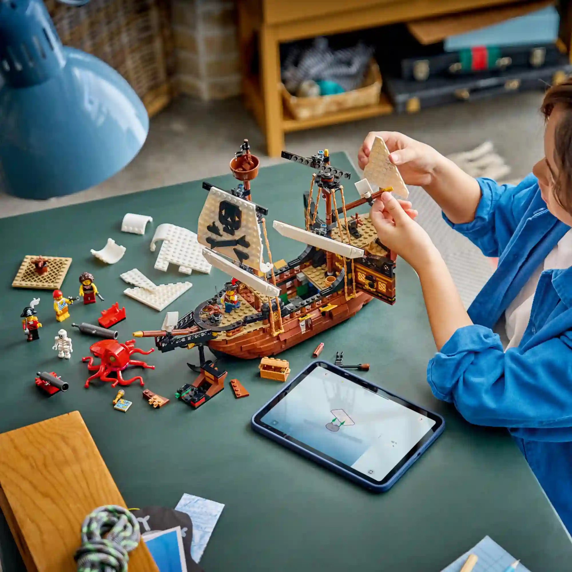 Gyermek építi a LEGO Creator Ikonikus kalózhajó (31387) modellt. Az asztalon szétszórt építőelemek, minifigurák és a LEGO Builder alkalmazást futtató táblagép látható, miközben a hajó vitorláját szereli fel.